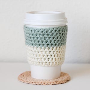 Pode incluir: Um cobertor de xícara de café de crochê em verde e branco com um descanso de xícara de crochê bege.