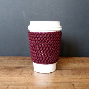 Könnte beinhalten: Ein bordeauxfarbener gehäkelter Kaffeetassenwärmer. Der Wärmer ist mit einem einfachen Stichmuster gearbeitet und für eine Kaffeetasse in Standardgröße konzipiert.