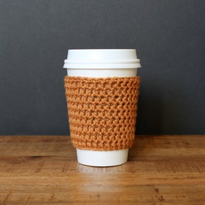 以下が含まれることがあります： 白いテイクアウトコーヒーカップに、茶色の編み物製のコースターが付いている。コースターはシンプルなステッチパターンで編まれている。