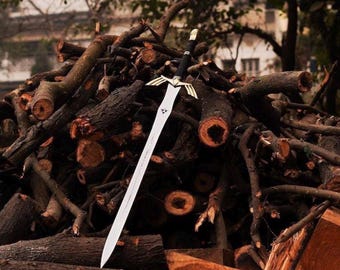 May include: A silver sword with a black handle and gold accents rests on a pile of cut logs. The blade is long and straight, with a pointed tip. The hilt features a crossguard with a decorative design.