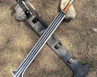 May include: Two crossed swords with black leather sheaths. The swords have silver blades with black stripes and wooden handles. The sheaths are black with straps and brass-colored rivets. The swords are lying on a bed of dry grass and pine needles.