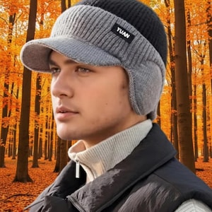 May include: A black, gray, and light gray knit hat with a visor and ear flaps. The hat has the word "YUAN" on a small label. The background is an autumn forest with orange and brown leaves.