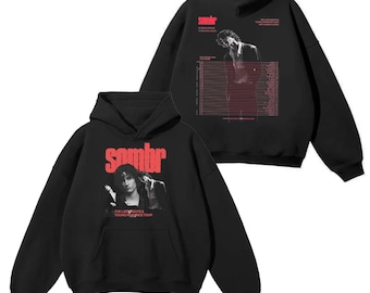 May include: Black hoodie with a front graphic featuring the word "sombr" in red, along with a black and white image of a person. The back of the hoodie also has a graphic with the word "sombr" and a black and white image.