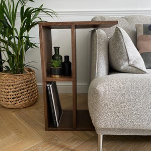 May include: A dark brown wooden side table with a built-in shelf, positioned next to a gray sofa. The shelf holds a small potted plant, two dark bottles, and a book. A large green plant in a woven basket is in the background.