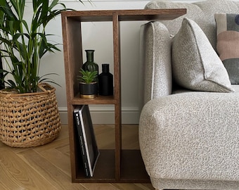 May include: A dark brown wooden side table with a built-in shelf, positioned next to a gray sofa. The shelf holds a small potted plant, two dark bottles, and a book. A large green plant in a woven basket is in the background.