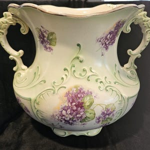 Pode incluir: Um jarro grande de cerâmica esbranquiçada com duas alças ornamentadas e uma borda ondulada. O jarro é decorado com desenhos florais de lilás roxo e detalhes pintados em verde. As alças têm um design escultural e decorativo.