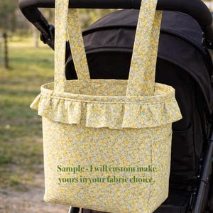 May include: A yellow floral fabric stroller bag with two top handles and a ruffled trim along the upper edge. The bag features the printed text: Sample - I will custom make yours in your fabric choice. The bag is attached to a black stroller frame.
