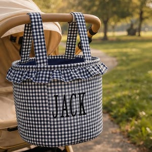 May include: A navy blue and white gingham fabric stroller basket featuring two matching handles, a ruffled top edge, and the name JACK embroidered in black capital letters on the front, hanging from a beige stroller handle.