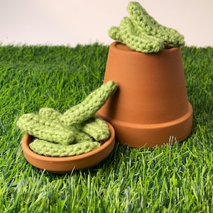 May include: A terracotta plant pot and small dish contain green crocheted cactus pieces. The cactus pieces are a light green colour and appear handmade. The items are set on artificial green grass.