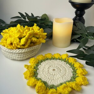 May include: A crocheted sunflower coaster with yellow petals, a white centre, and green accents. A matching crocheted bowl filled with yellow flowers sits nearby. A candle and greenery are in the background.