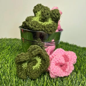May include: A small, silver bucket filled with green and pink crocheted flowers. Two green flowers and one pink flower are outside the bucket, resting on a bed of artificial green grass. The flowers have a textured appearance.