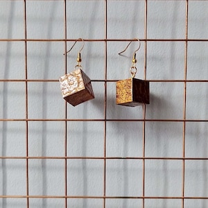 Könnte beinhalten: Ein Paar Ohrhänger mit würfelförmigen Anhängern, die eine dunkelbraune Holzmaserung und goldfarbene metallische Blattakzente auf den Flächen aufweisen. Die Ohrringe sind an goldfarbenen Metallhaken für durchstochene Ohren befestigt.