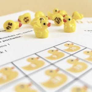 May include: A children's game with yellow plastic duck figurines, each with a red beak and a number. The game includes a matching card with duck illustrations and numbers. The ducks are small, likely under 5 cm in length.