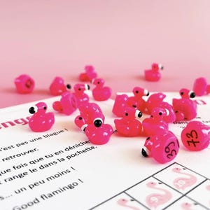 Può includere: Una collezione di piccoli pezzi di gioco rosa brillante a forma di fenicottero, con occhi bianchi e neri, sparsi su una carta da gioco bianca con illustrazioni di fenicotteri rosa. Lo sfondo è rosa pieno.