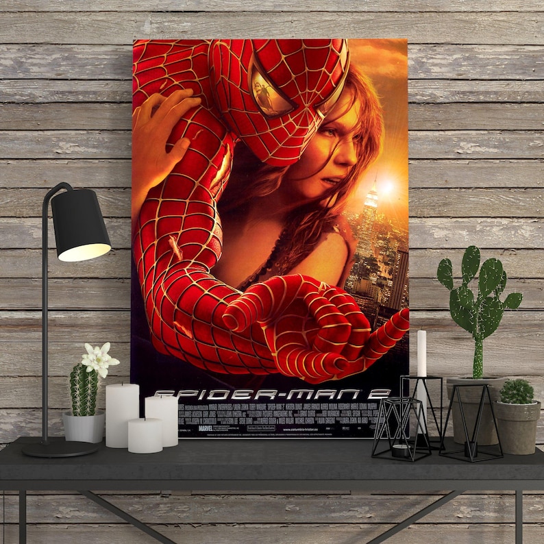 May include: A framed movie poster for Spider-Man 2. The poster features Spider-Man in a red and black suit embracing a woman. The background shows a cityscape and a warm, orange sky. The title is at the bottom.