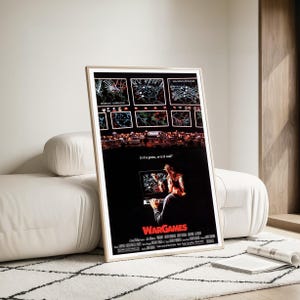 Puede incluir: Póster enmarcado de la película "WarGames". El póster presenta un fondo oscuro con imágenes de pantallas de ordenador que muestran mapas y datos. El título "WarGames" está en rojo, con el lema "¿Es un juego, o es real?"