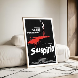 May include: A framed movie poster for Dario Argento's film, Suspiria. The poster features a black background with white text and a large red splatter design. The poster is in a light wood frame and is leaning against a white couch.