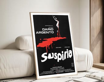 Puede incluir: Un póster enmarcado de la película Suspiria de Dario Argento. El póster presenta un fondo negro con texto blanco y un gran diseño de salpicaduras rojas. El póster está en un marco de madera clara y está apoyado contra un sofá blanco.
