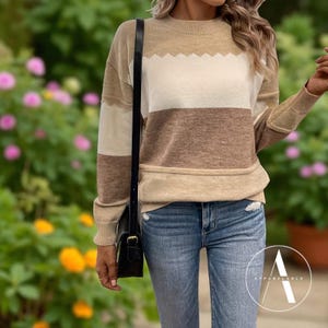 Puede incluir: Un suéter a rayas beige, blanco y marrón con cuello redondo. El suéter tiene un diseño festoneado en la parte superior de la franja blanca. Las mangas y el dobladillo inferior son de color beige liso. La modelo lleva vaqueros azules y un bolso negro.