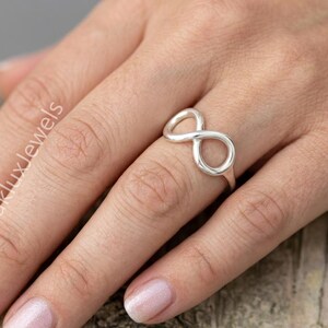Könnte beinhalten: Ein Ring aus poliertem Silber mit einem durchgehenden Unendlichkeitssymbol in der Mitte der Ringschiene. Das Metall hat eine glatte, reflektierende Oberfläche, die das Licht einfängt und eine klassische Schleifenform bildet.