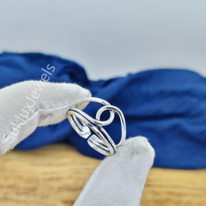 May include: A silver ring featuring an open, bypass-style design with two overlapping bands that form a circular loop at the top. The metal has a polished, reflective finish, and the band is adjustable with a visible gap at the bottom.