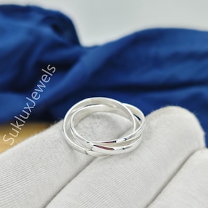 May include: A silver-coloured ring composed of three interlocking bands that create a continuous circular shape. The metal has a polished, reflective finish. The text SukluxJewels is visible in white lettering on the left side of the image.