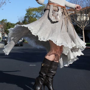 Puede incluir: Una mujer lleva una falda gris claro fluida con un cinturón de cuero marrón y detalles de flecos. Lleva botas de cuero marrón de tacón alto.