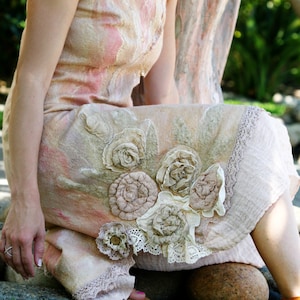 Op de afbeelding: Een vrouw draagt een lange, vloeiende, lichtroze jurk met bloemversieringen. De jurk heeft een kantversiering en is gemaakt van een zachte, vloeiende stof. De vrouw zit met gekruiste benen op een rots.