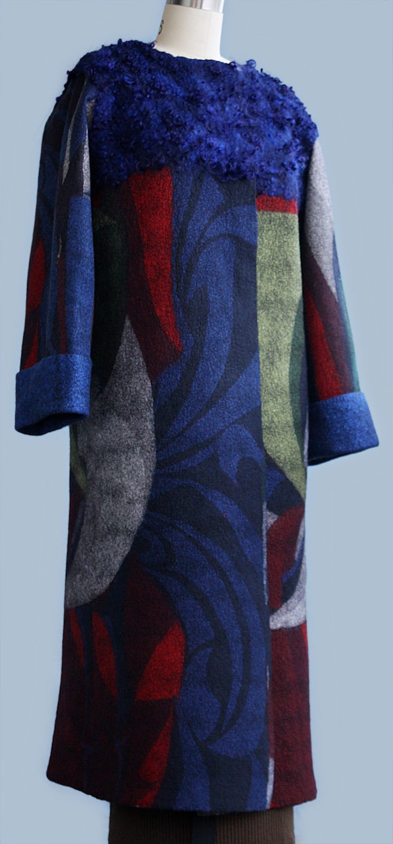 May include: A long, patterned coat with a blue, red, green, and gray design. The coat features a textured blue collar and cuffs. The coat is displayed on a mannequin.