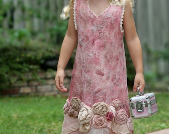 May include: A young girl wearing a pink dress with floral embellishments and a pink flower in her hair. She is holding a small pink and silver case.