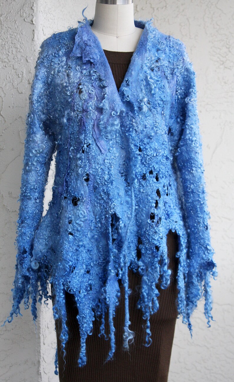 May include: A light blue, felted wool jacket with a unique, textured appearance. The jacket features a V-neck and long sleeves, with a fringed hemline. The wool has a curly, almost cloud-like texture, and the jacket is displayed on a mannequin.