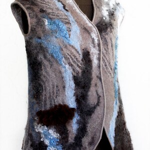 May include: A sleeveless vest with a unique, textured design. The vest features a mix of gray, blue, and brown colors, with a felted wool appearance. The design includes abstract patterns and a V-neck. The vest is displayed on a mannequin.