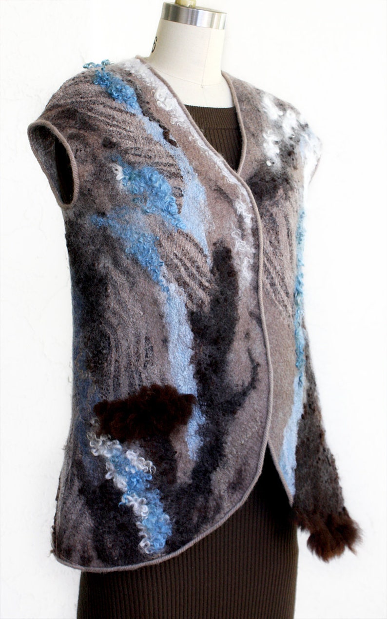 May include: A sleeveless vest with a unique, textured design. The vest features a mix of gray, blue, and brown colors, with a felted wool appearance. The design includes abstract patterns and a V-neck. The vest is displayed on a mannequin.