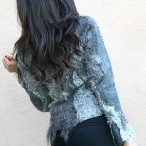 May include: A textured, gray jacket with a unique, fringed hemline. The jacket has a light blue hue and a detailed, textured design. The sleeves have a ruffled trim. The person is wearing black pants.