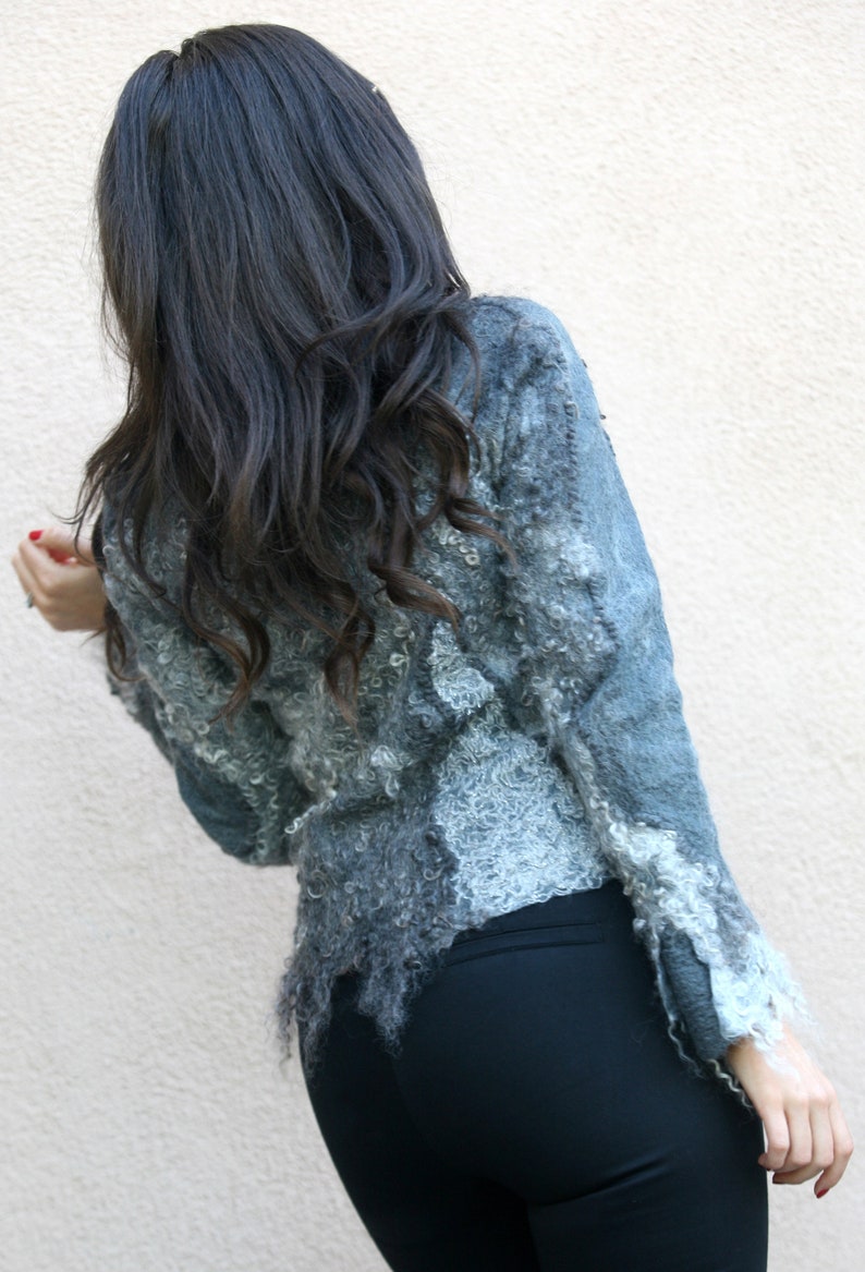 May include: A textured, gray jacket with a unique, fringed hemline. The jacket has a light blue hue and a detailed, textured design. The sleeves have a ruffled trim. The person is wearing black pants.