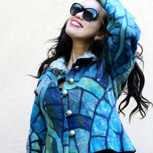 May include: A blue felted jacket with a mosaic-like pattern in shades of blue and green. The jacket has large silver buttons and a collar. The person is wearing black sunglasses and black pants.