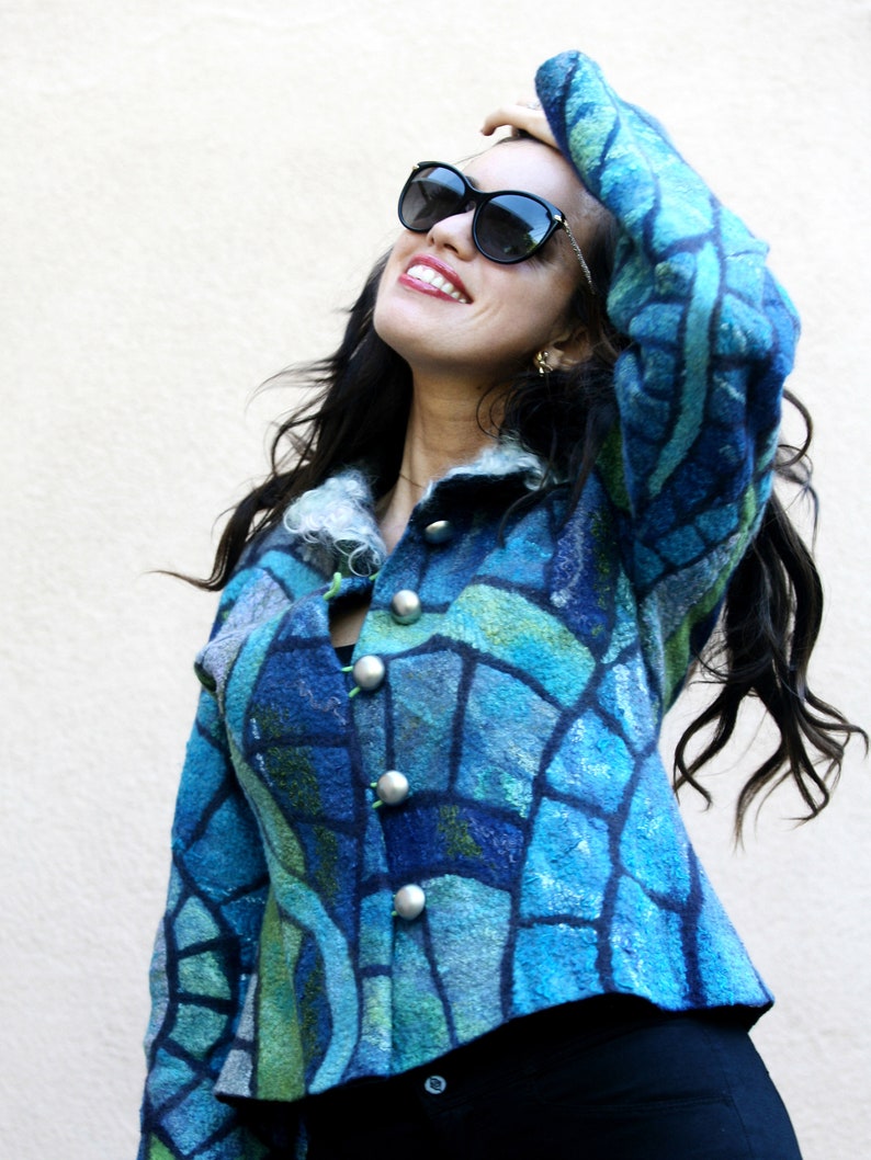 May include: A blue felted jacket with a mosaic-like pattern in shades of blue and green. The jacket has large silver buttons and a collar. The person is wearing black sunglasses and black pants.