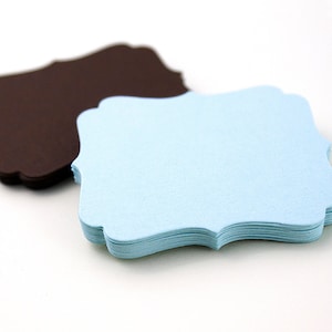 May include: A stack of light blue paper tags with scalloped edges, sitting on top of a stack of brown paper tags with scalloped edges.