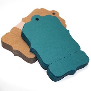 Peut inclure: Une pile d'&eacute;tiquettes cadeaux bleu sarcelle avec des bords festonn&eacute;s et un trou pour la ficelle. Les &eacute;tiquettes sont empil&eacute;es sur une pile d'&eacute;tiquettes en papier kraft brun.