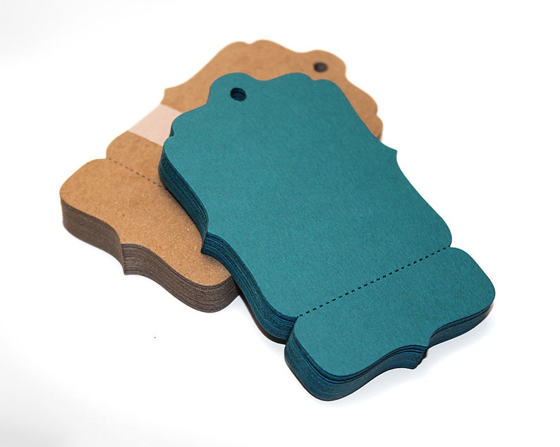 Peut inclure: Une pile d'&eacute;tiquettes cadeaux bleu sarcelle avec des bords festonn&eacute;s et un trou pour la ficelle. Les &eacute;tiquettes sont empil&eacute;es sur une pile d'&eacute;tiquettes en papier kraft brun.