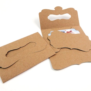 May include: Kraft paper gift card holders with decorative cut-out shapes. The holders are open, revealing a gift card inside one of them. The holders are made of brown cardboard and are designed to hold gift cards.