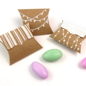 Puede incluir: Tres cajas de regalo de papel marr&oacute;n con dise&ntilde;os blancos. Los dise&ntilde;os incluyen rayas, una cadena de luces y una bandera. Tambi&eacute;n se muestran tres caramelos de almendras de color pastel.