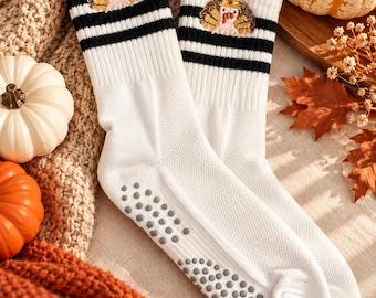 May include: A pair of white crew socks featuring two horizontal black stripes at the top and an embroidered turkey wearing a hat. The soles of the socks are covered in small, circular gray silicone grip dots for traction.