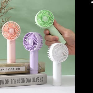 Peut inclure: Plusieurs ventilateurs portatifs aux couleurs pastel : rose, violet, vert et blanc. Chaque ventilateur a une tête circulaire avec un bouton central et une poignée. Un ventilateur est tenu. Les ventilateurs sont présentés sur des livres.