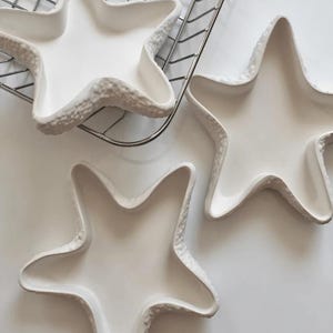 May include: Three white ceramic star-shaped dishes with textured edges. The dishes are arranged on a white surface, with one partially on a metal rack. The star dishes have a shallow bowl shape.