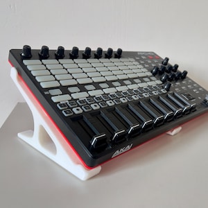 Op de afbeelding: Een zwarte en rode Akai APC-40 MKII MIDI-controller met witte knoppen en knoppen, rustend op een witte standaard. De controller heeft schuifregelaars en knoppen. De standaard is schuin voor optimaal gebruik.