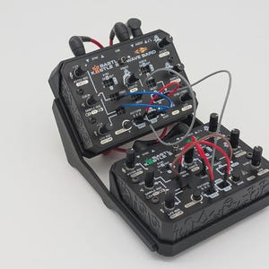May include: Black electronic music synthesizer with the text "BASTL INSTRUMENTS WAVE BARD" and various knobs, buttons, and ports. The device has multiple colored wires connected to it. The synthesizer is in a black case.