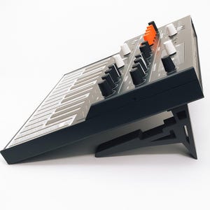 May include: A black synthesizer with white and orange knobs and a keyboard. The synthesizer is angled upward on a black stand, showcasing its controls and keys. The keys are white and gray.