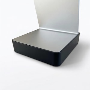 Pode incluir: Um suporte moderno e minimalista com um tampo prateado e uma base preta. O suporte tem uma base retangular e uma secção superior curva e aberta. O design é elegante e contemporâneo, adequado para exibir vários itens.