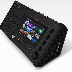 Może przedstawiać: Czarny Akai MPC Live centrum produkcji muzycznej z ekranem wyświetlającym różne obrazy. Urządzenie posiada liczne przyciski, pady i pokrętła. Tekst "AKAI MPC LIVE" jest widoczny na górze. Urządzenie jest ustawione pod kątem, pokazując jego bok z teksturowanym wzorem.